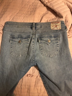 True Religion Joey ljusblå bootcut jeans - Säljer ett par ljusblå True Religion Joey jeans med bootcut passform. Klassiska jeansfickor med snygga sömmar och silverfärgade knappar. Märkespatch baktill och detaljerade ficklock på bakfickorna. Perfekta för dig som gillar ikoniska jeans med coola detaljer. Säljer dessa föratt de är för stora!!!❤️