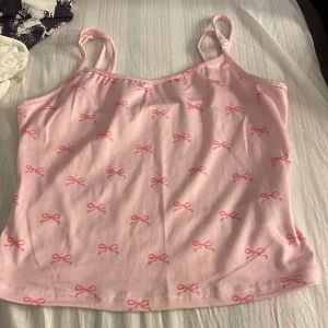 Rosa linnetopp med rosettprint - Supersöt ljusrosa linnetopp med justerbara axelband och små rosa rosetter som mönster över hela toppen. Mjuk och skön bomullskänsla, perfekt för varma dagar. Enkel och girly design som ger en gullig vibe.