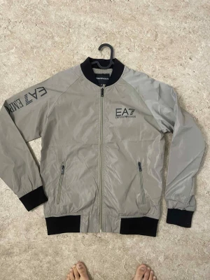 Grå bomberjacka från Emporio Armani - Snygg grå bomberjacka från Emporio Armani EA7 med svarta muddar och dragkedja framtill. Jackan har tryckt logga på bröstet och ärmen, samt två fickor med dragkedja. Perfekt för dig som vill ha en stilren och sportig look.