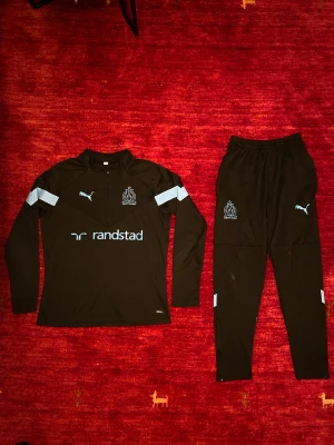 Marseille Tracksuit - Säljer ett stilrent och funktionellt träningsset från Puma i samarbete med Olympique de Marseille. Setet består av långärmad träningströja samt matchande träningsbyxor i svart med ljusblå detaljer. Klubbemblem, Puma-logotyper och sponsortryck ger ett autentiskt och sportigt uttryck.