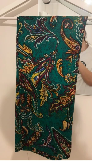 Grön långkjol med färgstarkt mönster - Säljer en grön långkjol med elastisk midja och fickor. Kjolen har ett färgstarkt paisley-inspirerat mönster i gult, lila, blått och rött. Materialet känns lätt och flowy, perfekt för dig som gillar att sticka ut med coola prints.