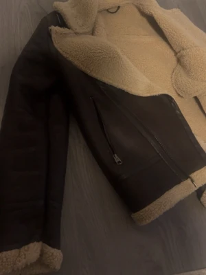 Brun teddyjacka med dragkedja - Snygg brun teddyjacka med krage och beige fluffigt foder. Jackan har två fickor med dragkedja och detaljer i svart skinnimitation. Perfekt för kalla dagar och har en loose passform. Fodret syns vid krage, ärmslut och nederkant för extra cozy vibe.