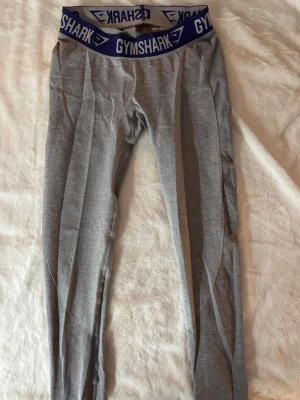 Grå Gymshark leggings  - Gråa leggings från Gymshark med bred blå resår i midjan där loggan syns. Tight passform och stretchigt material. Med detaljer på bakdelen.