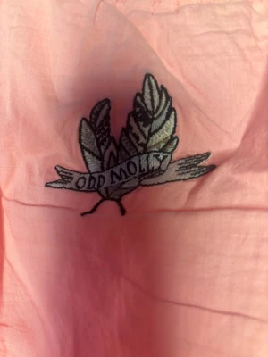 Rosa Odd Molly linnetopp med spets - Supersöt rosa linnetopp från Odd Molly med justerbara axelband, vit spetskant vid halsringning och nederkant samt en liten rosett framtill. Broderad Odd Molly-logga på ryggen och tygmärke vid nederkant. Perfekt för dig som gillar romantisk och feminin stil.