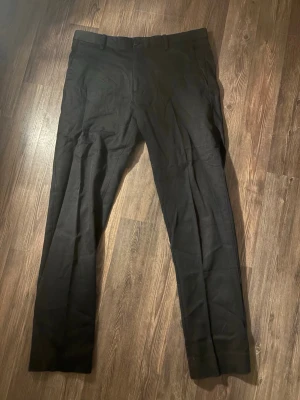 Svarta kostymbyxor från Riley - Snygga svarta kostymbyxor från Riley med klassisk rak passform och bälteshällor. Byxorna har en stilren look och är tillverkade i ett mjukt bomullsmaterial som känns bekvämt hela dagen. Perfekta för dig som gillar en clean och dressad stil.