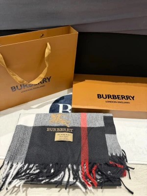 Burberry gray/black scarf -Unisex - Exklusiv väst från Burberry i 100% kashmir med klassiskt rutigt mönster i svart, grått, vitt och en röd detalj. Västen har fransar nedtill och en broderad Burberry-logga i guld. Perfekt för dig som vill ha en lyxig och tidlös look.