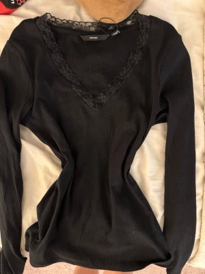 Svart ribbad långärmad topp med spets - Svart långärmad topp från Vero Moda med ribbad struktur och v-ringad hals. Halsringningen är dekorerad med snygg spetskant som ger en extra detalj. Modellen är figurnära och stretchig, perfekt till jeans eller kjol.