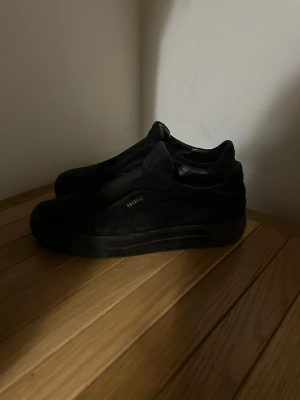 ARIGATOS SKOR - Säljer ett par helsvarta sneakers från ARIGATO med minimalistisk design och diskret logga i guld på sidan. Skorna har en låg siluett, platt sula och är tillverkade i mocka för en stilren look. Perfekta för dig som gillar clean och enkel stil.