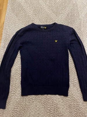Marinblå stickad tröja Lyle & Scott - Snygg marinblå stickad tröja från Lyle & Scott med klassiskt kabelmönster och gul logga på bröstet. Rund hals och ribbade muddar vid ärmslut och nederkant. Perfekt för dig som gillar stilrena och tidlösa plagg. Passar dig som är 158-167 cm lång