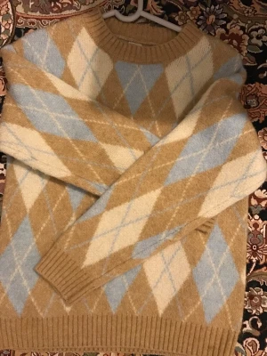 Beige och blå rutig stickad tröja - Säljer en mysig stickad tröja med rutigt argylemönster i beige, ljusblått och vitt. Tröjan har rund hals, ribbade muddar och långärmad passform. Perfekt för dig som gillar klassisk stil med en modern twist.