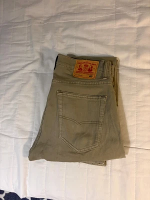 Beige jeansbyxor från Grant - Snygga beige jeansbyxor från Grant med klassisk femficksdesign och raka ben. Byxorna har en tydlig patch bak i midjan och är tillverkade i slitstark bomull. Perfekt för dig som gillar enkel och stilren look.