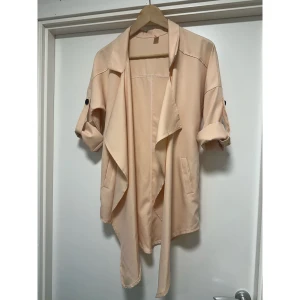 Beige kofta / jacka med uppvikbara ärmar - Beige / cremefärgad / ljust persikofärgad kofta / kavaj med långa ärmar som kan rullas upp och fästas med en knapp. Öppen framsida med en draperad krage och två dekorativa fickor fram. Lika fin öppen som med ett skärp i midjan. Storlek L/XL.   Hämtas i Stockholm eller skickas mot porto.