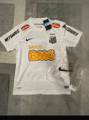 Vit Santos Neymar Jr matchtröja Nike - Santos FC matchtröja med Neymar Jr och nummer 11 på ryggen. Vit med svarta och orange tryck, klubbmärke, sponsorer och Nike-logga. Tillverkad i lätt polyester med Dri-FIT-teknik för skön känsla. Perfekt för dig som älskar fotboll och vill ha en ikonisk tröja.