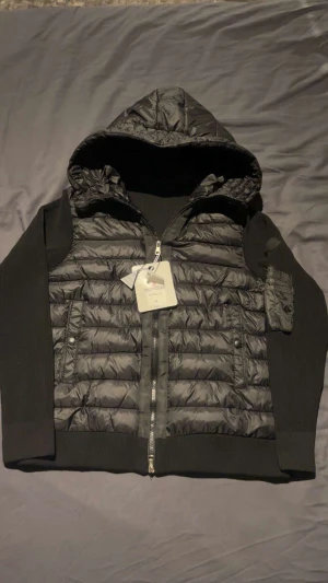 Svart Moncler pufferjacka med huva - Svart pufferjacka från Moncler med huva och snygga stickade ärmar. Jackan har quiltad front, dragkedja, och en praktisk ficka med zip på ärmen. Moncler-logga på ärmen och coolt innerfoder med serietryck. Perfekt för dig som gillar streetstyle och exklusiva märken.
