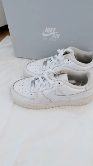 Nike Air Force 1 vita sneakers - Klassiska Nike Air Force 1 sneakers i helvitt skinn med perforerad tå och rund sula. Lågt skaft, snörning och ikonisk swoosh på sidan. Perfekt för dig som gillar clean och tidlös streetstil. 