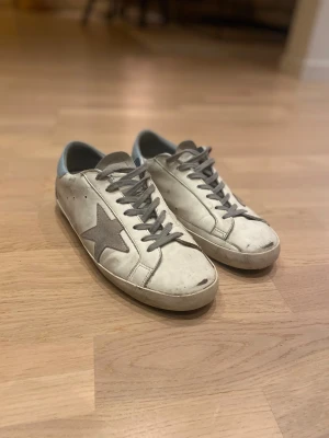 Golden Goose vita sneakers med stjärna - Golden Goose sneakers i vitt skinn med grå mockadetaljer och en stor grå stjärna på sidan. Skorna har grå snörning, ljusblå häl och platt sula. Klassisk rund tå och slitna detaljer för en cool, urban look.