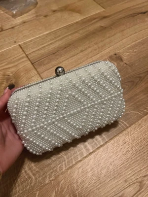 Vit pärlbeströdd kuvertväska - Snygg kuvertväska i vit färg med pärlbeströdd yta i geometriskt mönster. Väskan har ett metallspänne upptill och insidan är fodrad med svart tyg. Perfekt för att lyfta din outfit med lite extra bling och stil.