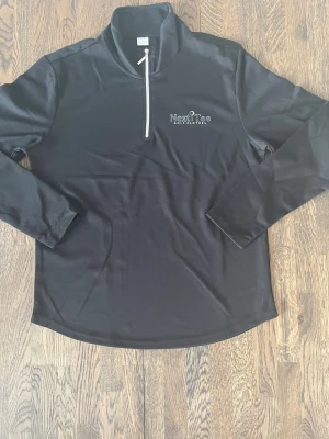 Svart golftröja Next Tee - Svart långärmad tröja från Next Tee Golf Clothes med vit dragkedja och diskret logga på bröstet. Tillverkad i 100% polyester med sportig känsla och stilren design. Perfekt för golf eller chill dagar. Snygg och enkel att matcha med andra plagg.
