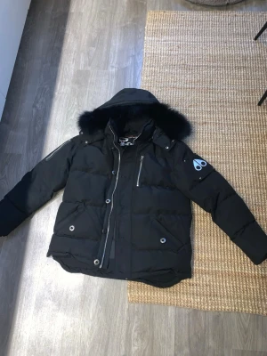 Svart pufferjacka Moose Knuckles L - Säljer en svart pufferjacka från Moose Knuckles i storlek L. Jackan har fluffig svart päls på huvan, flera silvriga dragkedjor och knappar, samt logga på ärmen. Fodrad med dun och har en cool, chunky look som är perfekt för kalla dagar. Den är dock rätt liten i storleken så jag tycker att den passar en person som har storlek M jag är 178cm lång och väger 75kg och jag har annars M i allt och den passar bra 