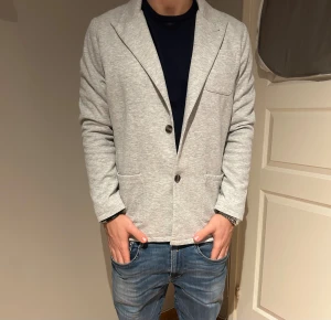 John Henric kavaj - Snygg ljusgrå kavaj med två knappar framtill och tre fickor. Tillverkad i mjukt bomullsmaterial som ger en avslappnad men ändå stilren look. Perfekt för dig som vill ha en modern och enkel kavaj med skön passform.