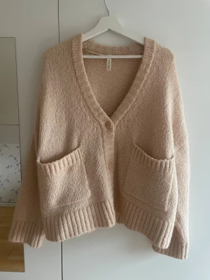 Beige oversized kofta från kay/day - Mysig beige kofta från Kappahl i oversized modell. Tröjan har stora fickor framtill, ribbade muddar och knäpps med två knappar. Perfekt för lager på lager och chill vardagsstil. Stickad i mjukt material och har en djup v-ringning.