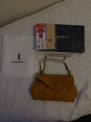 senapsgul quiltad axelväska från Yves Saint Laurent - Säljer en snygg gul axelväska från Yves Saint Laurent i mocka med quiltad design och guldfärgad kedja. Väskan har YSL-logga framtill och innerfack med dragkedja. Perfekt för dig som vill ha en lyxig och trendig accessoar.