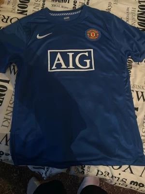 Blå Manchester United Ronaldo tröja - Säljer en blå Manchester United matchtröja med Ronaldo 7 på ryggen, tillverkad av Nike med FITDRY-teknologi. Tröjan har AIG-logga på bröstet, klubbmärke och vita detaljer. Perfekt för dig som älskar fotboll och Cristiano Ronaldo.