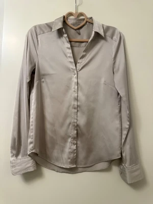 Champagnefärgad satinblus med knappar - Snygg champagnefärgad skjorta i satin med klassisk krage och långa ärmar. Skjortan har en slät yta, knäppning framtill och breda manschetter. Perfekt för dig som gillar en stilren och elegant look.