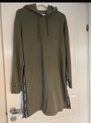 Olivgrön hoodieklänning Tommy Hilfiger - Olivgrön hoodieklänning från Tommy Hilfiger med långa ärmar och huva. Sidorna har snygga band med logotypen i vitt, rött och marinblått. Klänningen är i mjuk bomull och har en relaxed, loose passform. Perfekt för en chill och sportig look.