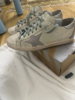 Golden Goose Superstar sneakers GGDB - Golden Goose Superstar sneakers i ljusbeige skinn med grå stjärndetalj på sidan och silverfärgad häl. Skorna har gröna snören, platt sula och rund tå. Märkets logga syns på hälen och insidan. Snyggt slitna detaljer och klassisk streetstil.