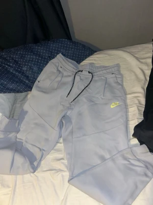 Baby blue Nike Tech byxor - Baby blue Nike Tech mjukisbyxor med gul logga på benet. Byxorna har svarta snören med vita detaljer i midjan och elastisk resår. Tillverkade i mjukt bomullsmaterial, har sidofickor och en avslappnad passform som är perfekt för chill eller träning. Köpta för runt 800-1000 kr. Pris kan diskuteras! 