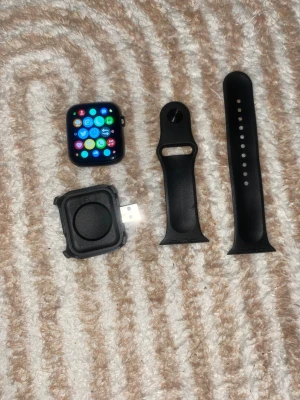Apple Watch (okänd modell) med svart armband och laddare - Apple Watch med svart armband och USB-laddare. Skärmen är igång och visar appar, inga synliga repor eller skador. Armbandet och laddaren ingår. Perfekt för dig som vill ha en smartklocka med många funktioner.