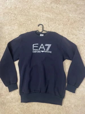 Mörkblå EA7 Emporio Armani sweatshirt - Snygg mörkblå sweatshirt från EA7 Emporio Armani med stor logotyp i silver på bröstet. Tröjan har rund hals, långa ärmar och ribbade muddar vid ärmslut och nederkant. Perfekt för dig som gillar stilrena och exklusiva märken.