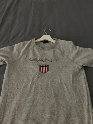Grå sweatshirt från GANT  - Säljer en grå riktigt fräsch sweatshirt från GANT med broderad logotyp och sköld på bröstet. Tröjan har rund halsringning och långa ärmar. Tillverkad i mjuk bomullsmix, perfekt för en chill och stilren look. Inga synliga defekter!  