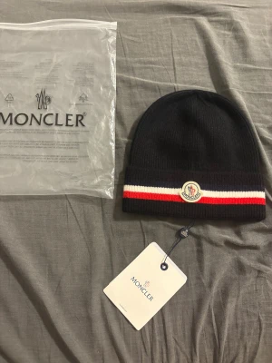 Moncler svart mössa med röd-vit rand - Svart ribbstickad mössa från Moncler med klassisk logga framtill och en snygg rand i rött och vitt längs kanten. Mössan är tillverkad i mjuk ull och har en clean, stilren look som passar perfekt till vinteroutfits. Taggar och originalpåse medföljer.