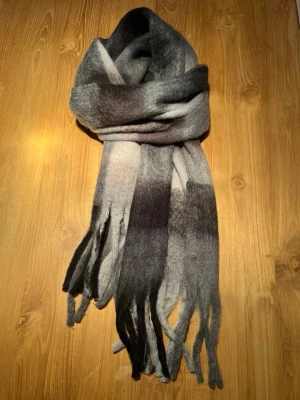 Grå och svart rutig ullhalsduk - Snygg halsduk i mjuk ull med rutigt mönster i olika nyanser av grått och svart. Halsduken  har en klassisk rak form och fransar längs nederkanten som ger en cool touch. Perfekt att slänga över en hoodie eller tröja för extra stilpoäng.