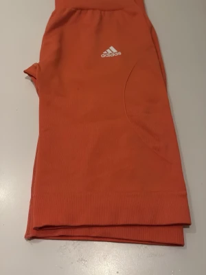 Orange träningsleggings från Adidas - Snygga orange leggings från Adidas med vit logga framtill. De har en tight passform och är gjorda i ett stretchigt syntetmaterial som passar perfekt för träning. Midjan är elastisk och benen är långa med ribbade muddar nedtill.