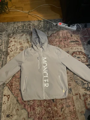 Grå vindjacka från Moncler med huva - Snygg grå vindjacka från Moncler med stor logotyp framtill, huva och tre dragkedjeförsedda fickor. Jackan har justerbar nederkant med snörning och Moncler-märke på ärmen. Perfekt för dig som gillar stilrena och exklusiva plagg.