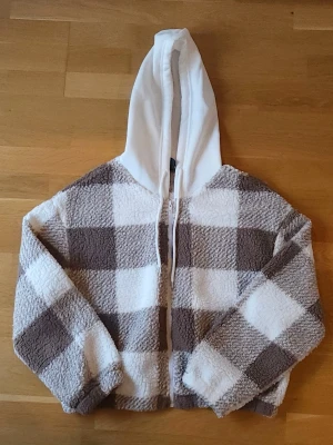 Rutig tröja - Supermysig tjocktröja i brun- och vitrutigt mönster. Tröjan har en vit huva med snörning och dragkedja framtill. Materialet är fluffigt och ger en chill vibe, perfekt för kyligare dagar.