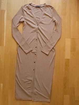 Beige klänning - Beige ribbad klänning med v-ringning och långa ärmar. Klänningen slutar ungefär mitt på vaden, har knappar hela vägen framtill och är tillverkad i ett mjukt, stretchigt material som sitter snyggt på kroppen. Perfekt för dig som gillar stilrena och bekväma plagg.