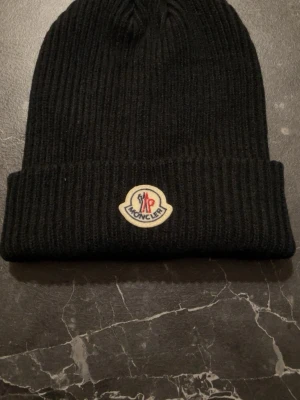 Moncler mössa - Snygg svart ribbstickad mössa från Moncler med klassisk logotyp framtill. Mössan är tillverkad i mjuk ull och har en uppvikt kant som ger en cool och enkel look. Perfekt för dig som vill ha en stilren accessoar med exklusiv känsla.