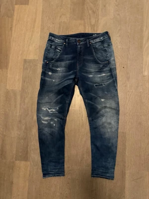 Blåa vintage jeans från diesel - Säljer ett par blå jeans som är från diesel, med coola slitningar och tvättade detaljer. Modellen är croppad med avsmalnande ben och har klassiska fem fickor. Jeansen har en mörk tvätt med ljusa partier och är tillverkade i jeansmaterial. Perfekt för dig som gillar en avslappnad streetstil.