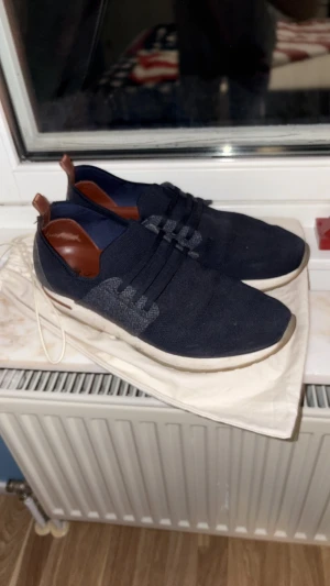 Mörkblå sneakers från Loro Piana - Snygga mörkblå sneakers från Loro Piana med stickad ovandel och detaljer i brunt skinn. Skorna har rund tå, platt sula och snörning framtill. Insidan är fodrad med brunt läder och hälen har en mockadetalj. Perfekt för dig som gillar stilrena och bekväma sneakers.