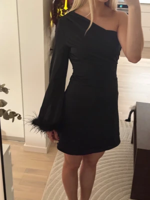 Slutsåld nyårsklänning - Snygg svart kortklänning från Feeling Friday i modellen Lovisa Dress, perfekt till nyår. Aldrig använd, prislapp kvar (nypris 1259kr + tull). Klänningen har en oneshoulder-design med lång ärm och fjäderdetaljer vid ärmslutet. På deras hemsida står det att det är ”high quality satin”.