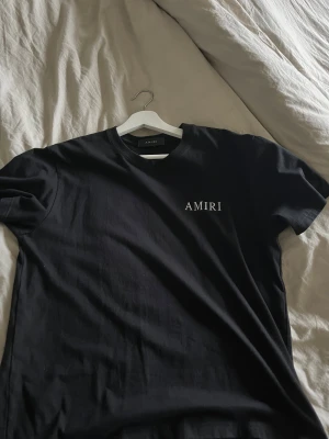 Svart AMIRI t-shirt med ängeltryck - Svart t-shirt från AMIRI med vit logotyp på bröstet och stort konstnärligt tryck av en ängel på ryggen. Klassisk rund hals och korta ärmar. Materialet är mjuk bomull som känns skönt mot huden. Perfekt för dig som gillar streetwear med exklusiv vibe.