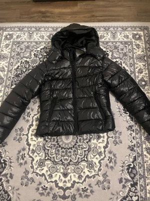 Svart pufferjacka från lager 157 L - Snygg svart pufferjacka från 157 i storlek L. Jackan har en glansig finish, huva med tryckknappar och två dragkedjeförsedda fickor framtill. Perfekt för kalla dagar och har en modern, quiltad look som är lätt att matcha med din stil. Haft på mig den max 2 gånger finns i Hudiksvall och posta