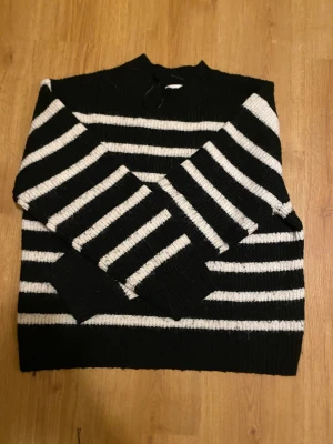 Svartvit randig stickad tröja - Säljer en svart och vit stickad tröja med breda horisontella ränder. Tröjan har långärmade ärmar och en högre hals med en liten knytning bak. Den är ribbstickad i muddar och nederkant, perfekt för dig som gillar klassisk och chill stil.
