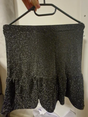 Glittrig svart volangkjol - Supersnygg svart kortkjol med glittriga detaljer och volangkant. Kjolen har en figurnära passform upptill och faller snyggt med volanger nedtill. Perfekt för dig som vill sticka ut med lite extra glitter. Materialet är mjukt och följsamt.