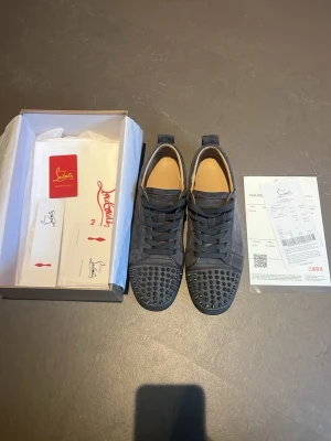 Christian Louboutin mörkgråa sneakers med spikar - Säljer ett par svarta sneakers från Christian Louboutin i mocka med nitar på tån. Skorna har snörning och klassisk rund tå. Kommer med originalkartong, dustbag och äkthetskort. Perfekta för dig som vill sticka ut med exklusiv streetstyle.