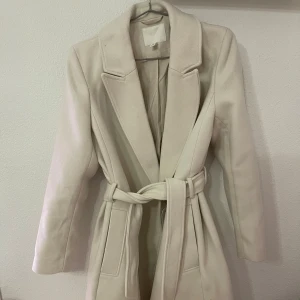 Beige kappa med bälte från H&M - Snygg beige kappa från H&M i storlek 36. Jackan har klassisk krage, långa ärmar och ett matchande bälte i midjan för en clean och stilren look. Perfekt för dig som gillar minimalistisk stil och vill ha en jacka som funkar till många outfits. Orginalpris 999kr